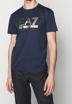 Man wearing a navy blue EA7 Emporio Armani t-shirt with gold logo lettering and black jeans, standing against a plain background.
