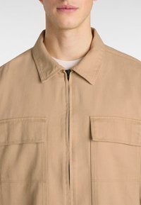 Tan, heavy-duty jacket featuring a zip closure, pointed collar, and two chest pockets. Fabric has a textured, durable appearance.
