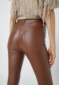 Leggings marron en similicuir avec une taille haute, deux poches arrière et un design élégant avec une texture lisse et brillante.