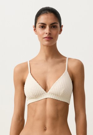 Femeie cu păr închis la culoare, purtând un sutien texturat alb, orientată frontal, pe un fundal simplu și deschis la culoare.