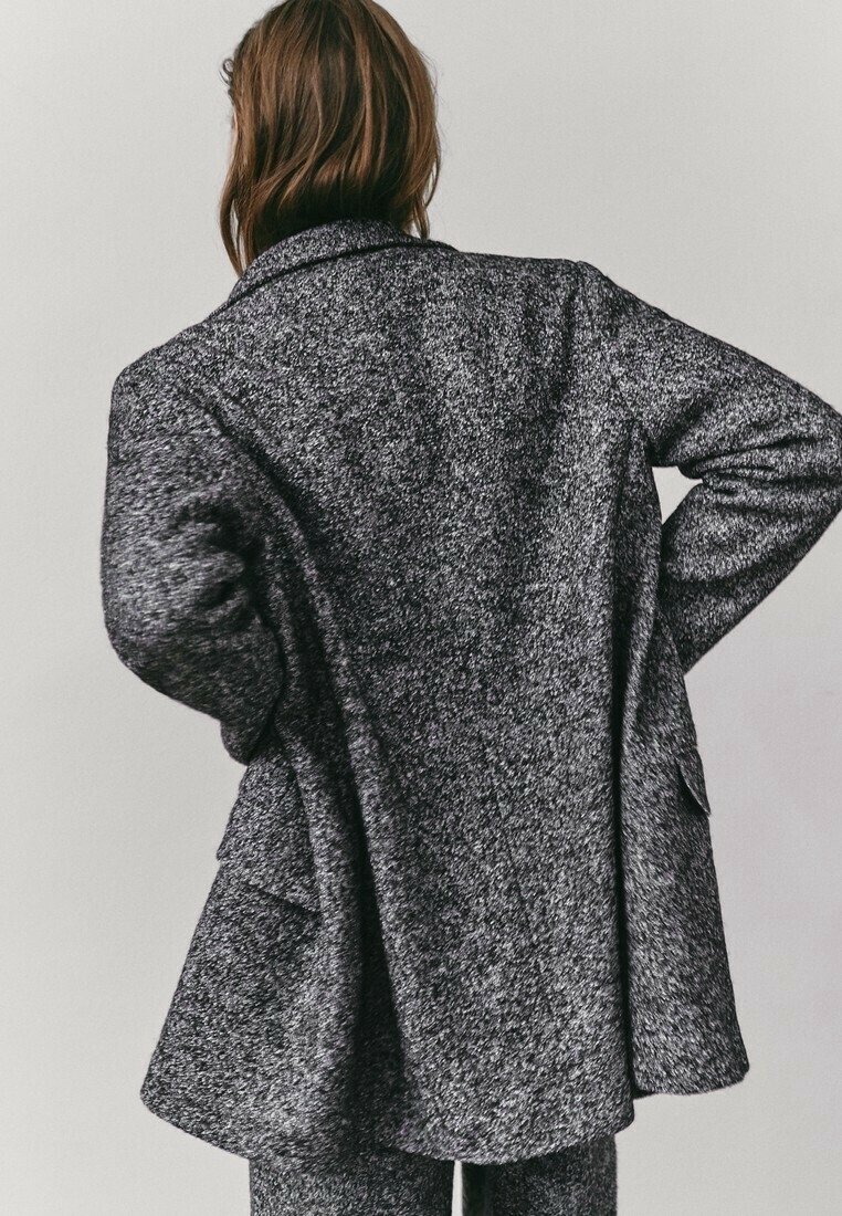 Femme portant un manteau gris texturé à manches longues et un pantalon assorti, debout les mains sur les hanches, de dos sur un fond uni.