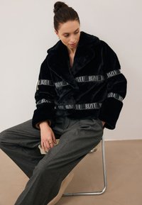 Czarna kurtka z futerka z kołnierzem typu lapel, z szerokim paskiem z logo "BIZUU". W połączeniu z szarymi spodniami w plisy i beżowym obuwiem.