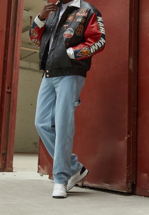 Osoba w czarno-czerwonej kurtce varsity z naszywkami, jasnoniebieskich luźnych jeansach i białych trampkach, stojąca przy czerwonych drzwiach.