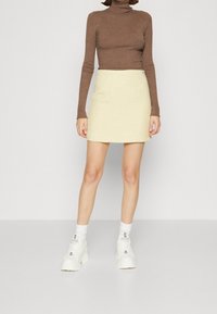 Bruine geribbelde turtleneck trui gecombineerd met een lichtgele gestructureerde minirok. Witte chunky sneakers en opgerolde witte sokken maken de look compleet.