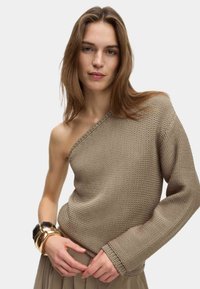 Pull en tricot beige avec une épaule dénudée. Tissu texturé, manche longue d'un côté, fini avec de délicates coutures.