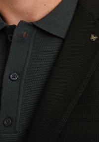 Donkergroene gemêleerde polo met een knoopsluiting, zwarte knopen en een klein gouden embleem op de kraag naast een zwarte jas.