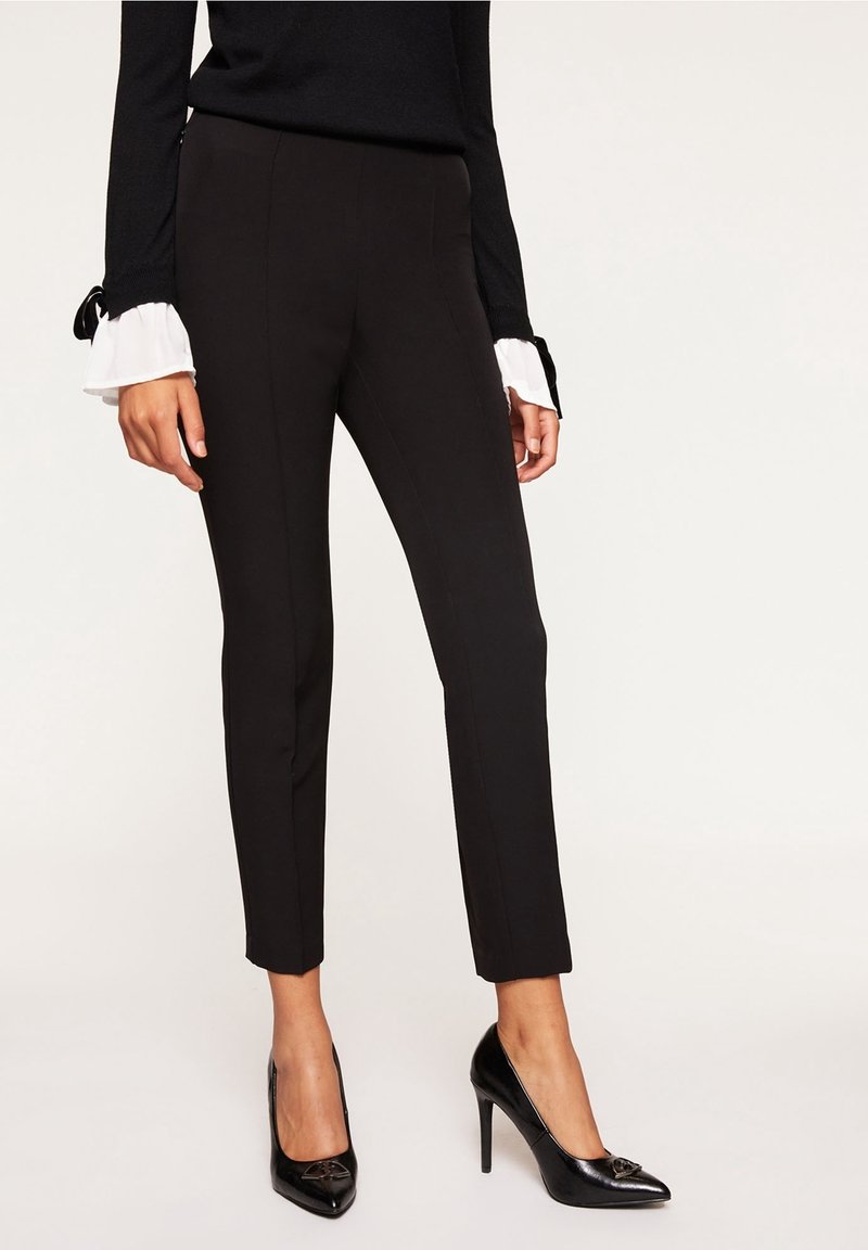 Femme portant un pantalon noir ajusté à la cheville, des escarpins noirs pointus et un haut noir avec des poignets à volants blancs, debout devant un fond uni.
