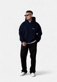 Sweatshirt à capuche bleu marine en tissu doux, avec une poche avant et un détail de logo. Associé à un pantalon noir et des baskets blanches.
