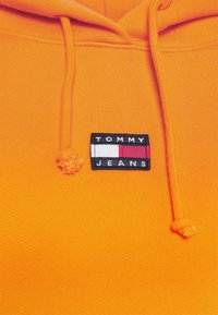 Sudadera con capucha naranja con cordones, que presenta un parche rectangular negro con la inscripción "TOMMY JEANS" y franjas gráficas en rojo, blanco y azul.