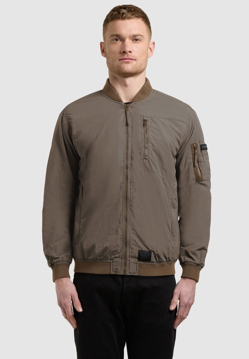 Homme portant une veste aviateur marron avec des poches zippées sur la poitrine et les manches, debout devant un fond clair uni.