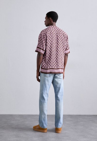 Homme de dos portant une chemise à manches courtes rose et brune à motifs, un jean bleu clair et des baskets en daim beige, se tenant sur un sol gris.