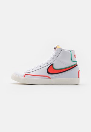 Sneaker Nike alta bianca con swoosh bordato in rosso e verde, lacci bianchi e suola beige chiaro su sfondo neutro.