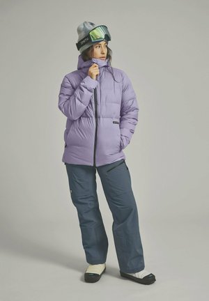 Purple puffer jacket with a high collar and zipper, paired with dark blue snow pants and white slide sandals; features goggles and a beanie.
