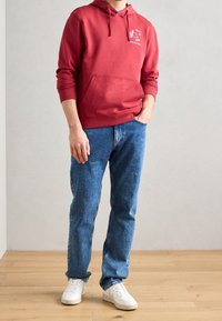 Felpa rossa in cotone con tasca frontale, caratterizzata da una grafica bianca. Indossata con jeans in denim blu e sneakers bianche.