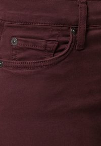 Los jeans de denim burdeos presentan una textura suave con un bolsillo frontal y acentos de hardware plateado. Las costuras alrededor de los bolsillos son visibles.