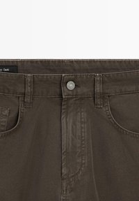 Pantalon marron foncé avec passants pour ceinture, fermeture par bouton métallique, poches avant et détails de coutures visibles au niveau de la taille.