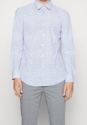 Homme portant une chemise blanche à manches longues boutonnée avec un motif vertical en tirets bleus et un pantalon gris clair.