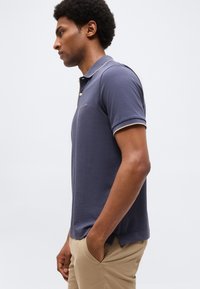 Mladý muž stojaci v profile, oblečený v námornícky modrej polokošeli a béžových nohaviciach, s rukami v vreckách, na jednoduchom pozadí.