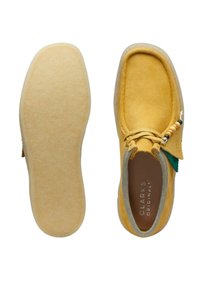 Gula mockaskor med en mjuk, texturerad ovandel, en ljusfärgad sula och en dekorativ snörning med träpärlor och en grön accent.