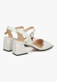 Witte hakken sandalen met een vierkante blokhak, open teen en kruisvormige banden met gesp. Gladde textuur en minimalistisch ontwerp.