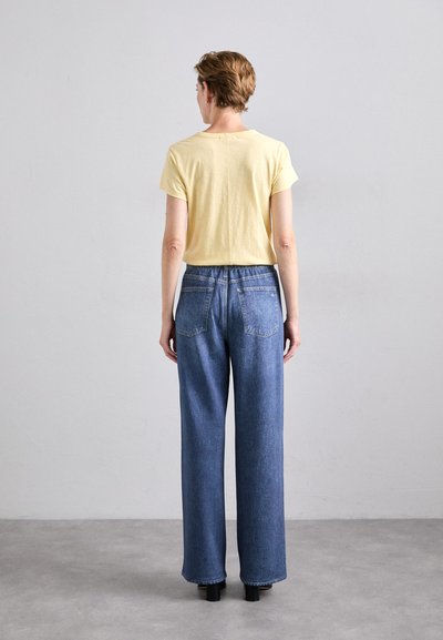 Gul kortermet t-skjorte kombinert med høyt liv, brede blå jeans med baklommer. Modellen har på seg svarte hæler.