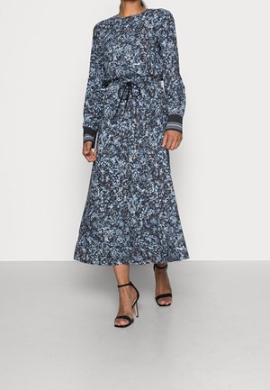 Žena v dlouhých rukávech, dlouhých až k kotníkům, květinových šatech s vázáním v pase, v černých sandálech na vysokém podpatku, kráčí vpřed.