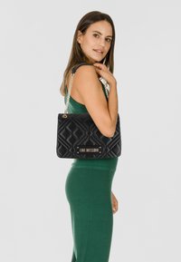 Borsa a tracolla nera trapuntata con catena argentata e placca logo dorata. Presenta una forma rettangolare e una superficie liscia.