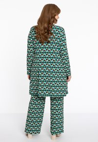 Grünes gemustertes Outfit, bestehend aus einer Tunika mit langen Ärmeln und verlängertem Saum sowie passenden weit geschnittenen Hosen, die beide geometrische Muster zeigen.