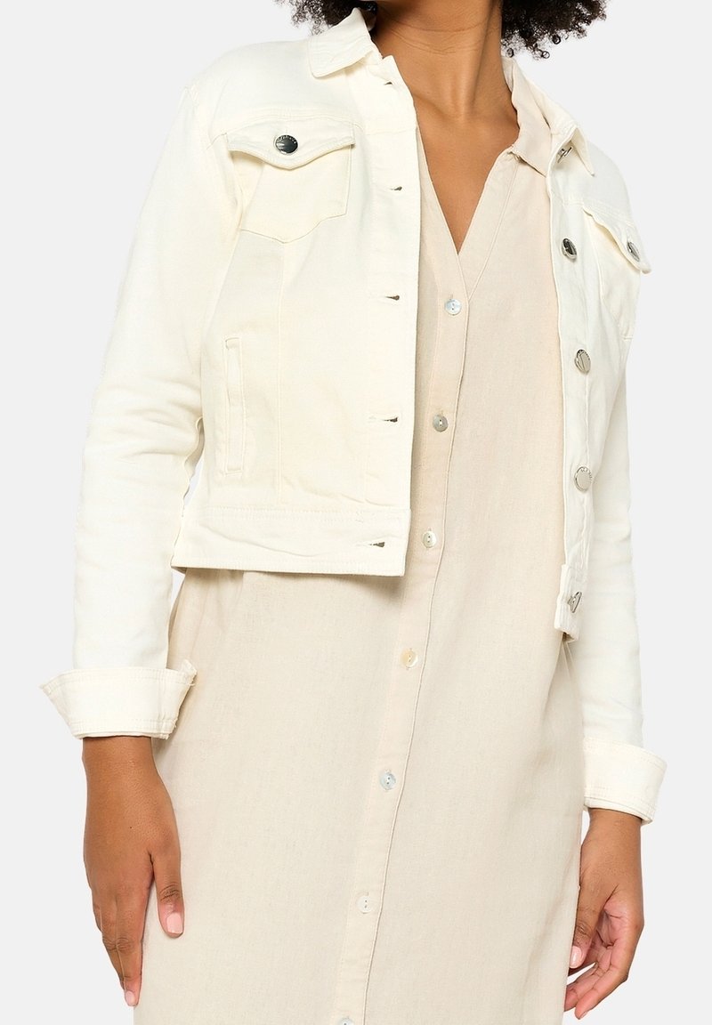 Veste en denim blanc avec des boutons en argent, comportant deux poches à l'avant et des manches retroussées, portée sur une robe beige à boutons.