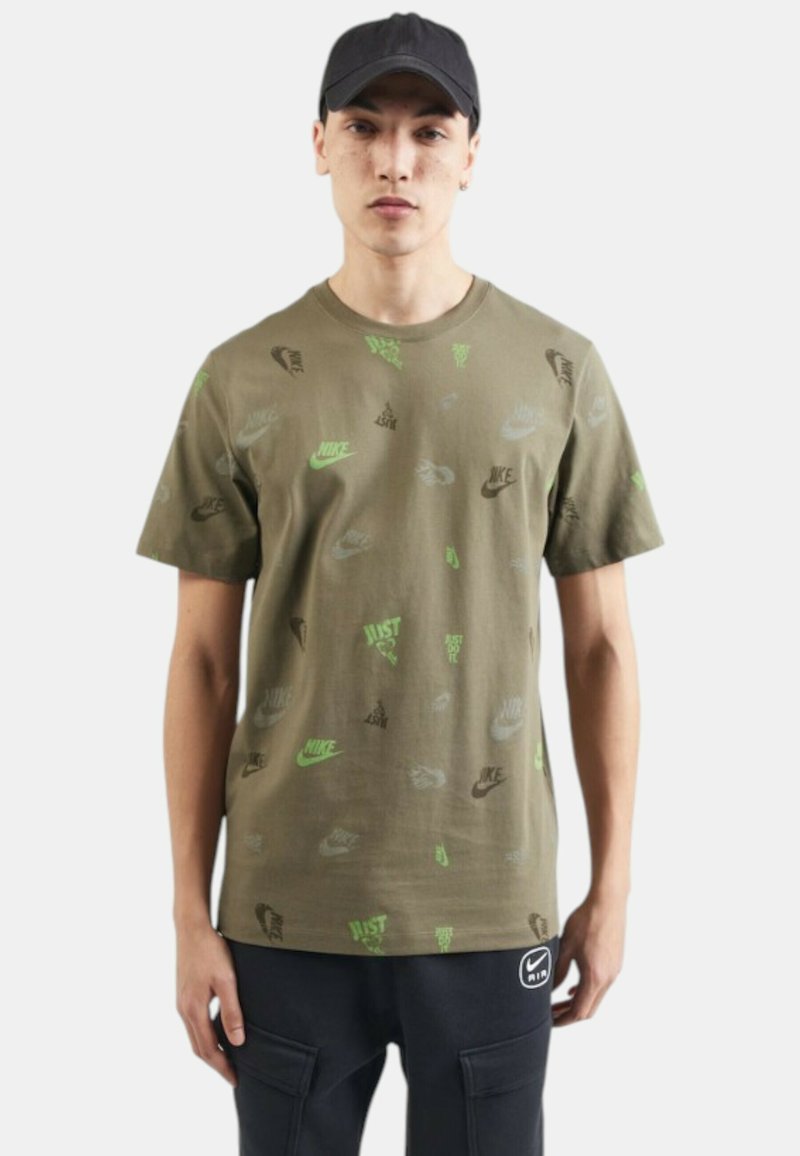 Camiseta de algodón verde oliva con logotipos de Nike dispersos en negro y verde neón. Mangas cortas, cuello redondo, ajuste relajado, diseño sencillo.
