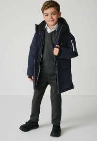 Chaqueta aislante azul marino con un bolsillo con cremallera, suéter gris y pantalones oscuros. Zapatos negros con cinta de velcro. El modelo sonríe a la cámara.
