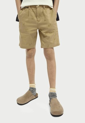 Beżowe szorty z delikatnym wzorem żagli, o luźnym kroju i podwiniętych nogawkach, zestawione z beżowymi zamszowymi chodakami i szarymi skarpetami z żółtymi akcentami.