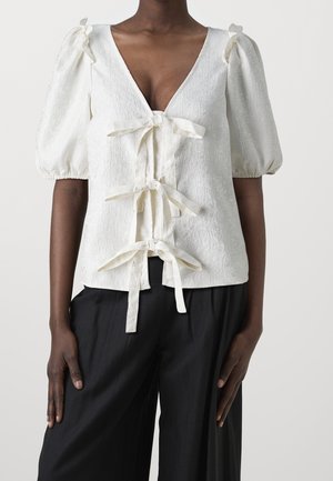 Une personne à la peau noire portant un blouson blanc à manches bouffantes avec trois nœuds devant et un pantalon noir ample, sur un fond uni.