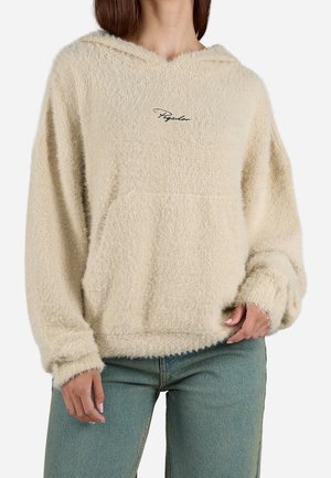 Beige gebreide trui met capuchon, voorzak en een cursief logo op de borst, gedragen met een blauwe spijkerbroek door een persoon met een medium huidtint.