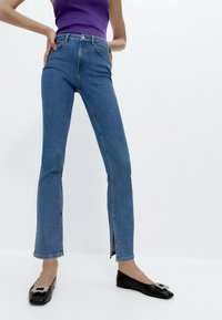 Ljusblå denimjeans med utsvängda ben och hög midja, med en liten slits i fållen. Matchas med svarta ballerinaskor prydda med en dekorerad rosett.