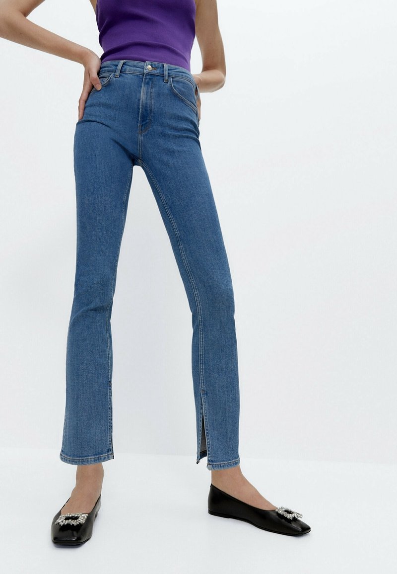 Ljusblå denimjeans med utsvängda ben och hög midja, med en liten slits i fållen. Matchas med svarta ballerinaskor prydda med en dekorerad rosett.