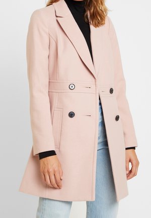 Femme portant un manteau en laine rose clair à double boutonnage sur un col roulé noir et un jean bleu clair, visible du menton jusqu'à mi-cuisse.