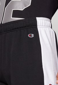 Black and white athletic pants with an elastic waistband, featuring a Champion logo in red and white on the thigh. Soft fabric texture.