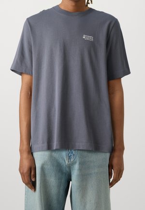 Graues Baumwoll-T-Shirt mit kurzen Ärmeln, Rundhalsausschnitt und kleinem weißen Logo auf der linken Brustseite. Kombiniert mit hellblauen Denim-Jeans.