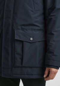 Marineblå isolert jakke med sidelommer, med trykknapplukking. Teksturen ser glatt ut og designet er funksjonelt.