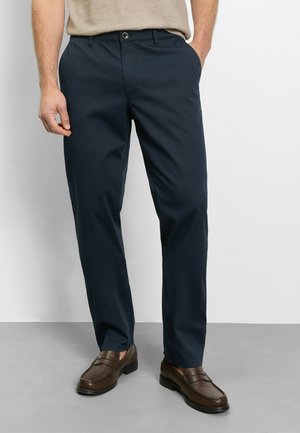 Man draagt marineblauwe broek met rechte pijpen en bruine leren loafers, staat met één hand in de zak tegen een effen achtergrond.