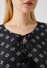 Μαύρη μπλούζα με σχέδιο από λευκά τετράγωνα, κλείσιμο με κορδόνι στη λαιμόκοψη και μαύρες φούντες. Μαλακή υφή με άνετη εφαρμογή.