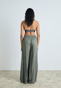 Svart Calvin Klein-märkt sportbehå och lösa, olivgröna vida byxor i mjukt tyg. Bakgrunden är ljusgrå.