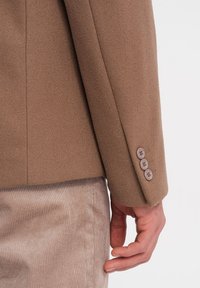 Bruine wollen blazer met een getailleerde pasvorm. Heeft een gladde textuur, drieknoopmanchetdetails en subtiele stiksels op de mouw.
