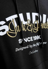 Mustalne kapuutsiga pusa, millel on suur valge ja kuldne trükk. Tekst sisaldab "STUDIO Jack & Jones" ja "Disainitud J&J meeskonna poolt." Reguleeritav kapuuts.