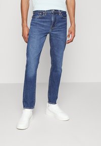 Jeans de mezclilla azul con corte recto, que cuentan con cinco bolsillos, costuras naranjas y un cierre clásico de botón. Llevados con zapatillas blancas.