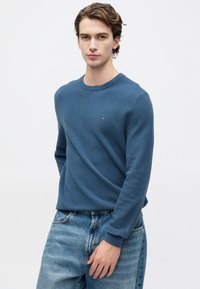 Jauns vīrietis, tērpts teksturētā zilā džemperī un izbalējušās zilās džinsās, stāv uz vienkārša gaiša fona.
