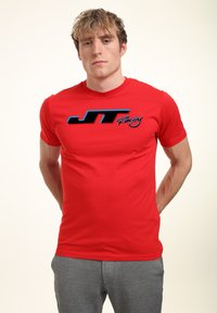 Červené tričko s krátkým rukávem vyrobené z bavlny s hladkou strukturou. Na přední straně se nachází výrazné logo "JT Racing" v modré a černé barvě.