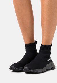 Black knit ankle sneakers with textured upper and flexible sole. Features logo detail on the side and a ribbed collar for a secure fit.
