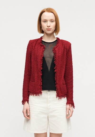 Veste rouge texturée avec détails en franges. Encolure ronde, devant ouvert et deux poches, portée sur un haut noir et un short crème.